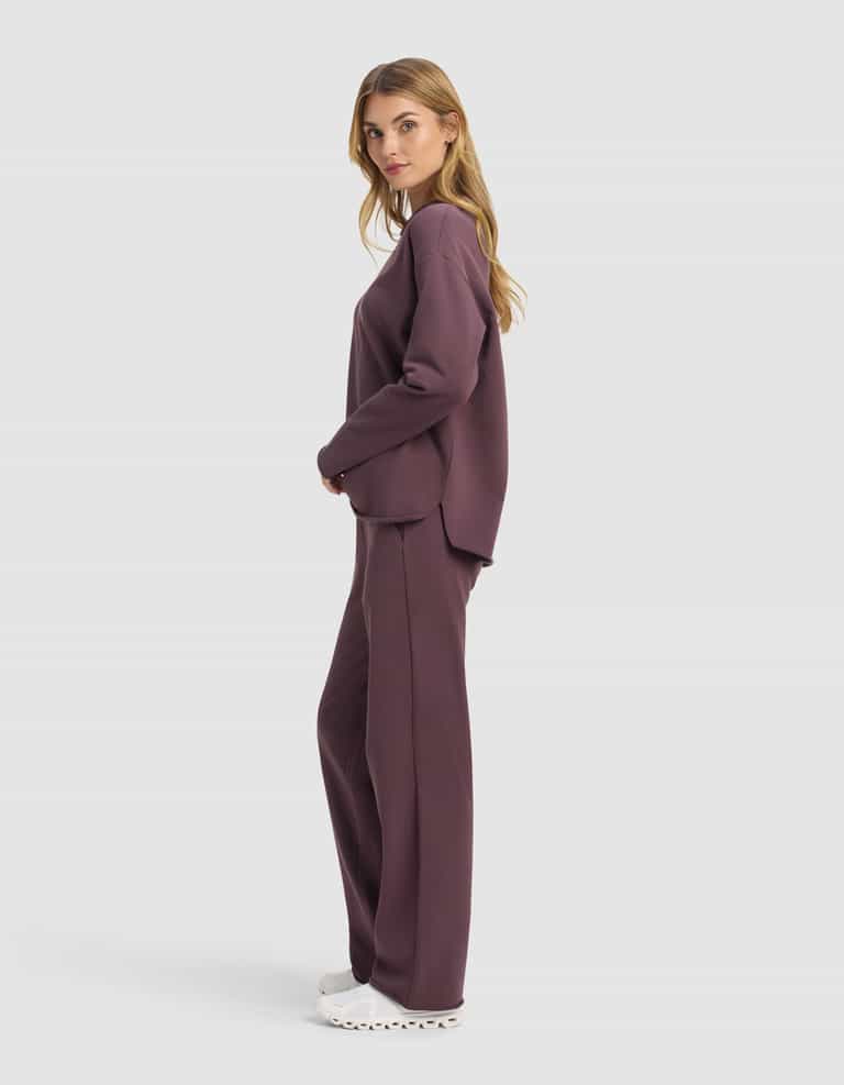 A woman stands in profile wearing the Cozy Earth Women's Chelsea Set—loose-fitting, long-sleeve dark mauve sweatshirt and matching pants—with white sneakers, set against a plain light background.|Color:Fig