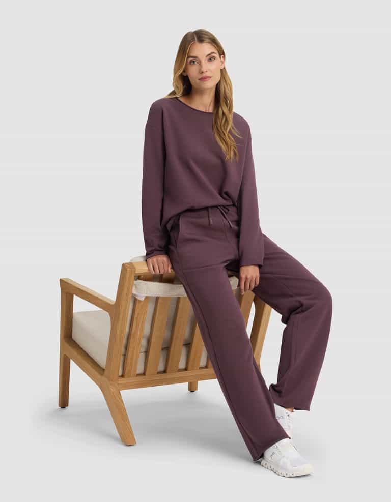 A woman wearing Cozy Earth's Women's Chelsea Pullover in dark mauve and matching pants sits on the armrest of a wooden chair with beige cushions, her long hair down and white sneakers on, looking calmly at the camera.|Color:Fig