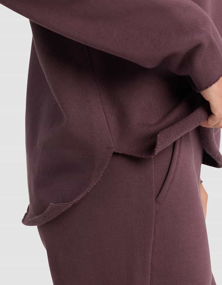 A close-up of a person wearing the Cozy Earth Women's Chelsea Pullover in maroon, featuring a hand partly in the pocket and a rounded, notched hem detail at the side.|Color:Fig