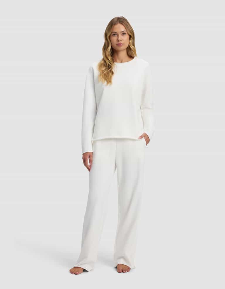 A woman with long blonde hair models the Cozy Earth Women's Chelsea Set—a loose-fitting, long-sleeve white top and matching pants—standing barefoot against a light gray background, gazing at the camera with a neutral expression.|Color:Bone
