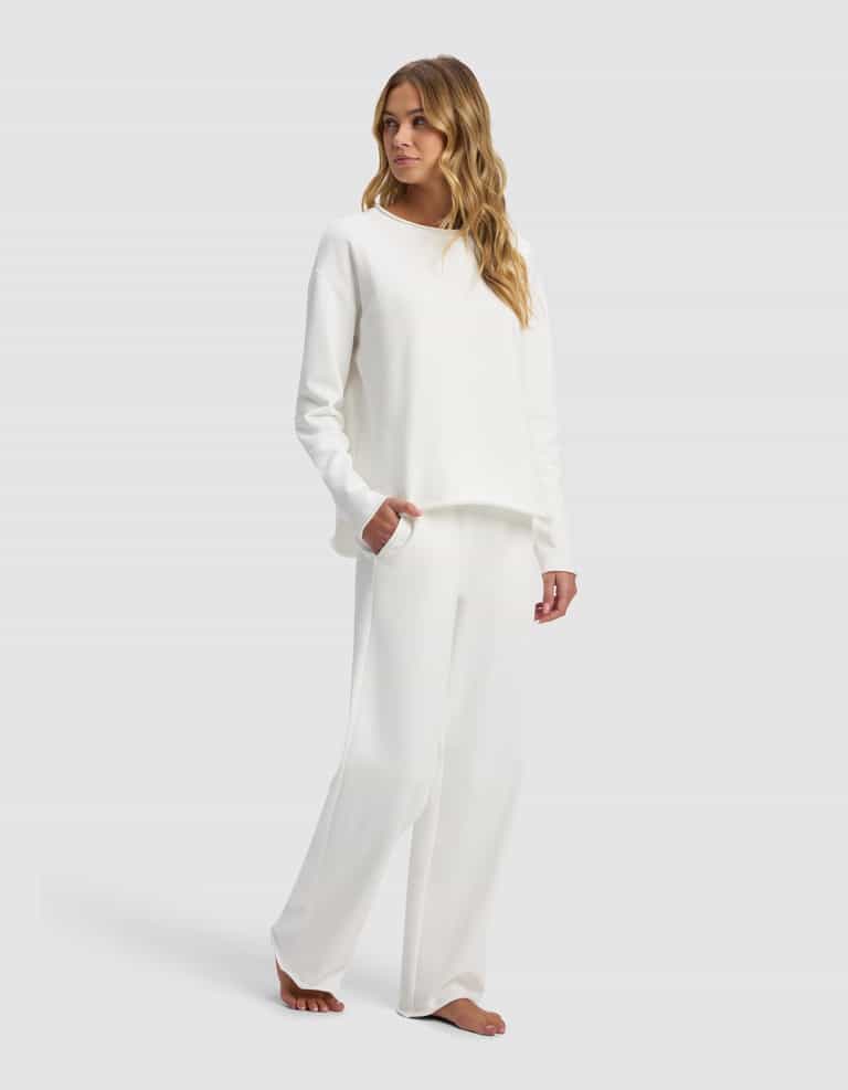 A woman with long wavy hair stands barefoot against a plain light background, wearing the Women's Chelsea Set by Cozy Earth—a loose-fitting, long-sleeve white top and matching wide-leg white pants.|Color:Bone