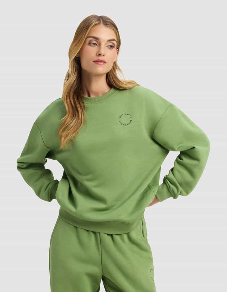 A woman with long blonde hair stands with her hands on her hips against a plain light gray background, wearing the Cozy Earth Women's Cityscape 2.0 Crewneck in a loose-fitting, light green style with matching sweatpants.|Color:Clover