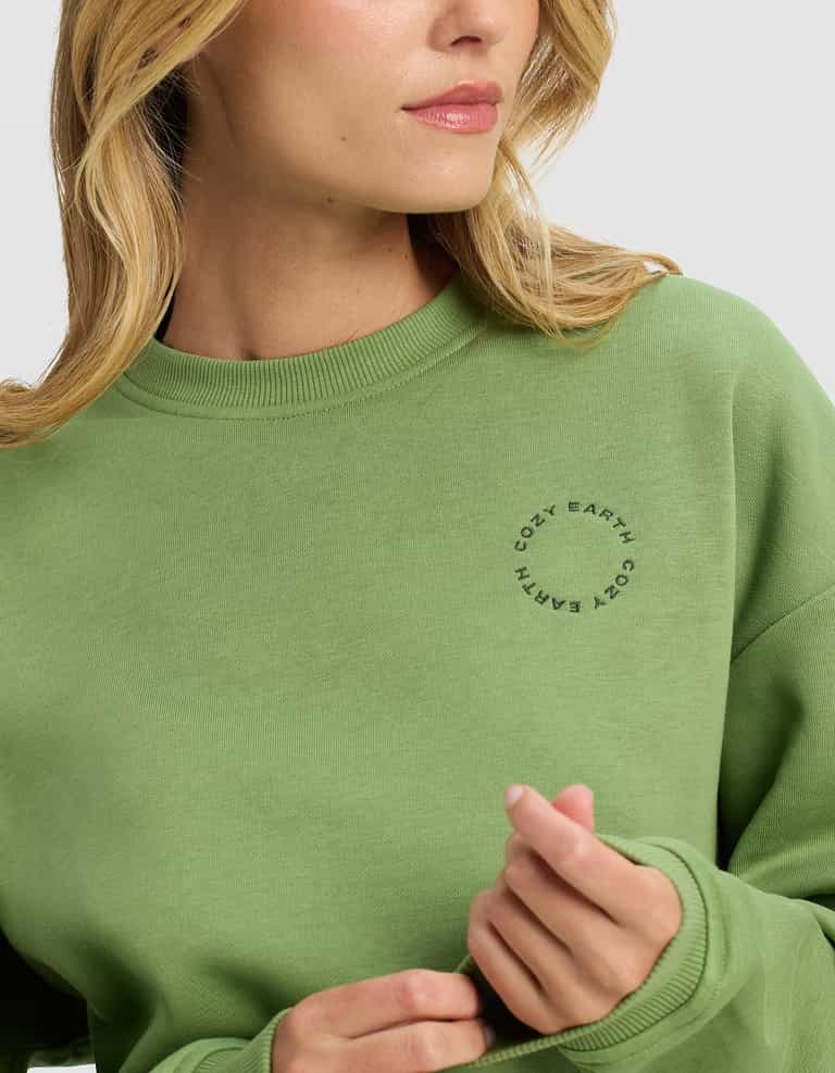 A woman wearing the Cozy Earth Women's Cityscape 2.0 Crewneck in light green poses against a plain background, facing slightly to the side with her hand near her collar.|Color:Clover
