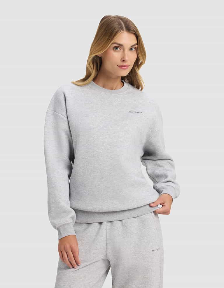 A woman with long blonde hair models the Cozy Earth Women's Cityscape 2.0 Crewneck in light gray, paired with matching sweatpants, standing with one hand in her pocket against a plain light background.|Color:Heather Grey