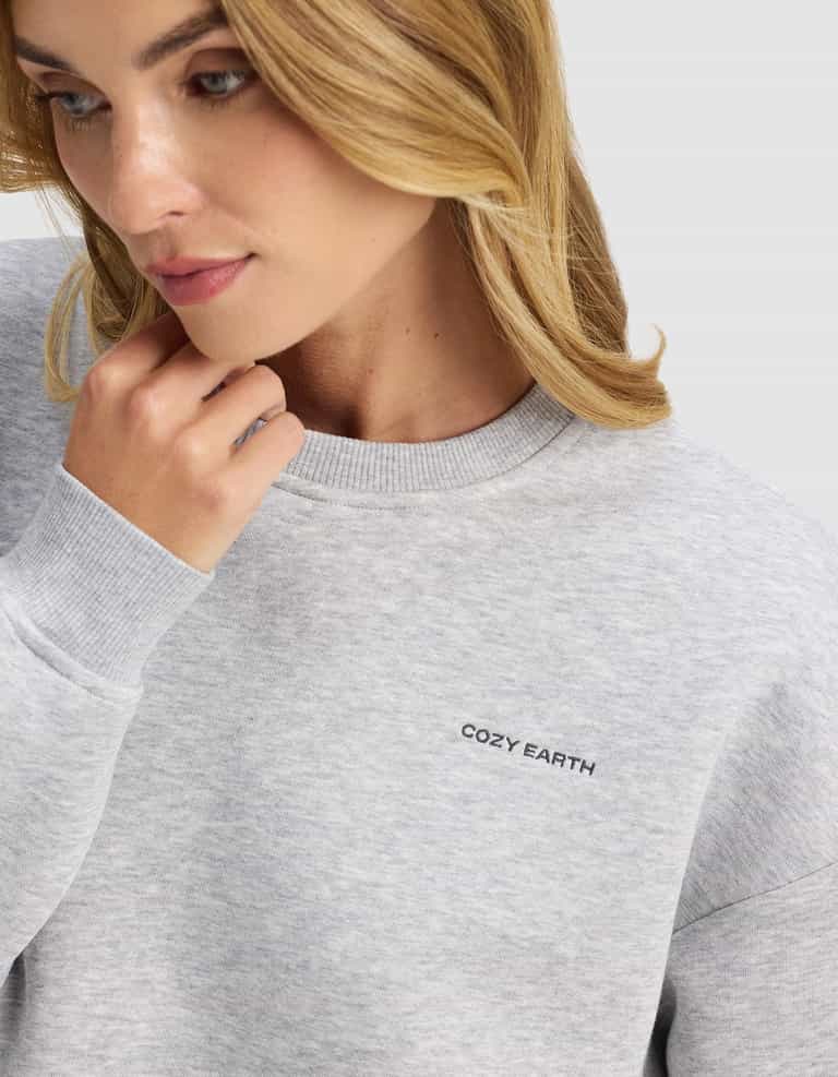 A woman with wavy blonde hair wears the Cozy Earth Women's Cityscape 2.0 Crewneck in light gray, featuring the brand embroidered on the chest, as she gazes down thoughtfully and touches her chin.|Color:Heather Grey