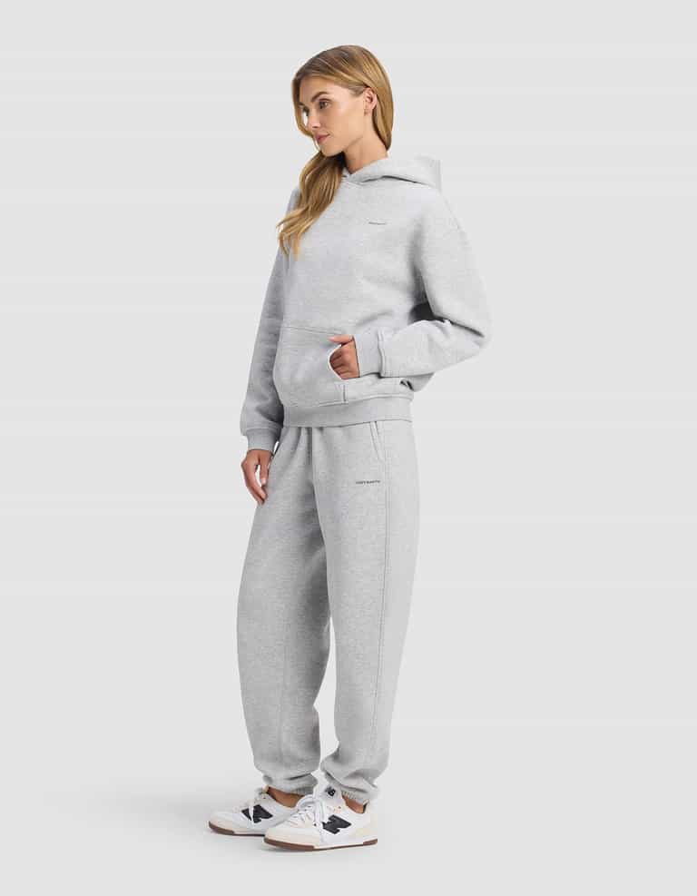 A woman with long blonde hair stands in a relaxed pose, hands in the front pocket, wearing Cozy Earth's Women's Cityscape 2.0 Hoodie & Sweatpant Set in light gray with white sneakers against a plain light gray background.|Color:Heather Grey