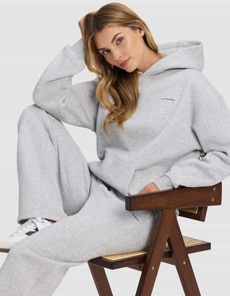 A woman with long blonde hair sits on a wooden chair, wearing the Cozy Earth Women's Cityscape 2.0 Hoodie in light gray, with matching sweatpants. She rests one arm on her head and gazes relaxingly at the camera.|Color:Heather Grey