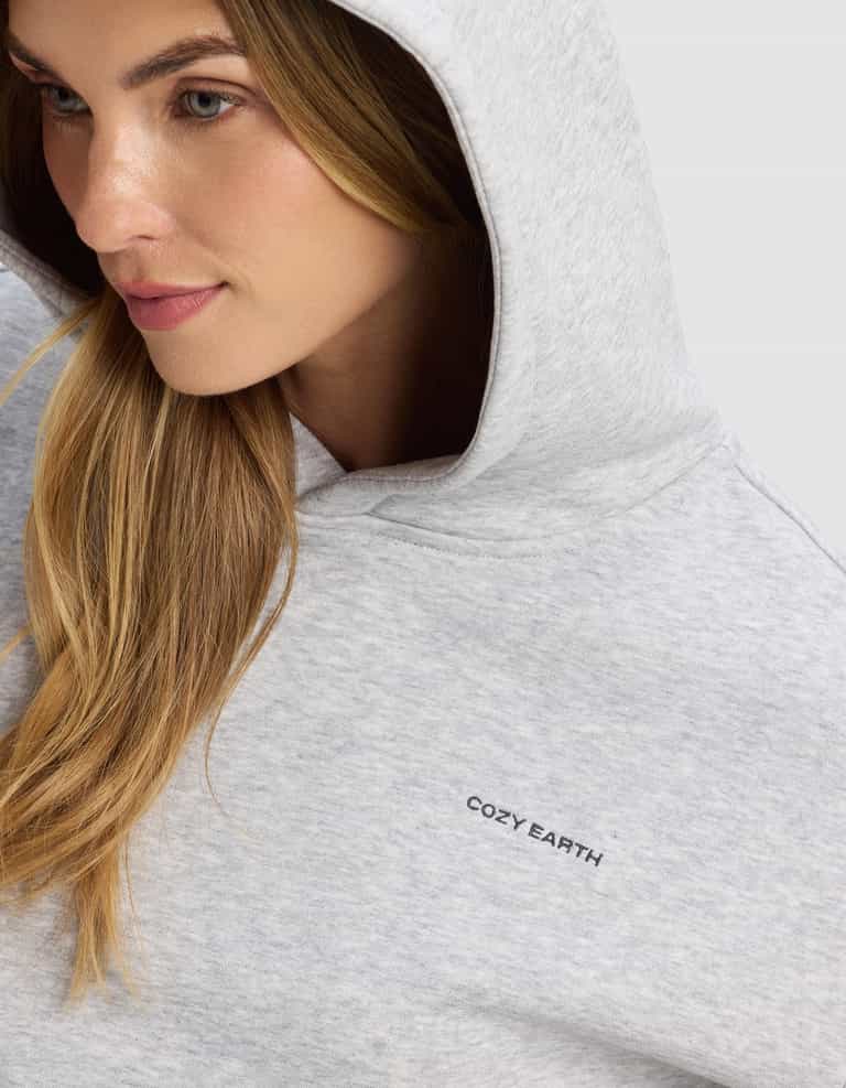 A woman with long blonde hair wears the Cozy Earth Women's Cityscape 2.0 Hoodie in light gray, featuring a small black "COZY EARTH" logo on the chest. She looks slightly to the side with a gentle smile.|Color:Heather Grey