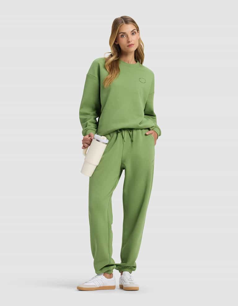 A woman models the Cozy Earth Women's Cityscape 2.0 Sweatpant in green with a matching sweatshirt, white sneakers, and a cream water bottle, posing with one hand in her pocket against a plain light background.|Color:Clover
