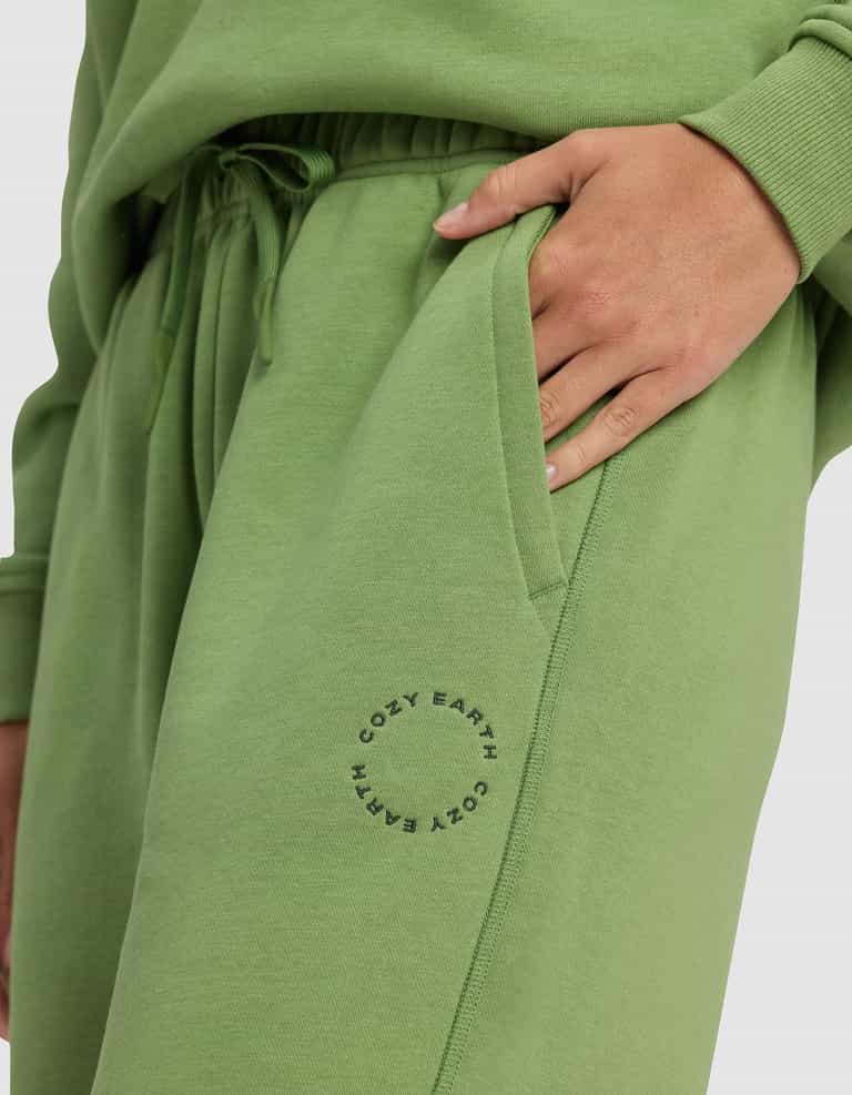 A person wears Cozy Earth's Women's Cityscape 2.0 Sweatpant in light green with a matching sweatshirt, one hand in their pocket. "COZY EARTH" is printed in a circle near the sweatpant’s pocket.|Color:Clover