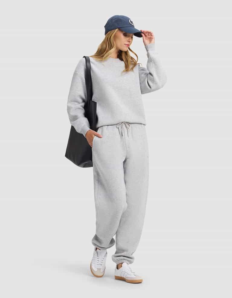 A woman in Cozy Earth's Cityscape 2.0 Sweatpant, a gray sweatshirt, white sneakers, navy blue cap, and carrying a large black tote poses against a plain background with one hand in her pocket and the other adjusting her cap.|Color:Heather Grey