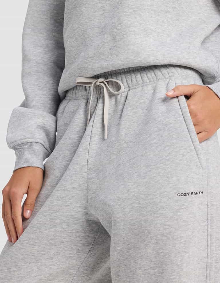 A person wears light gray Women's Cityscape 2.0 Sweatpants by Cozy Earth, featuring an elastic waistband with drawstring, one hand in a pocket, and "COZY EARTH" printed on the side.|Color:Heather Grey