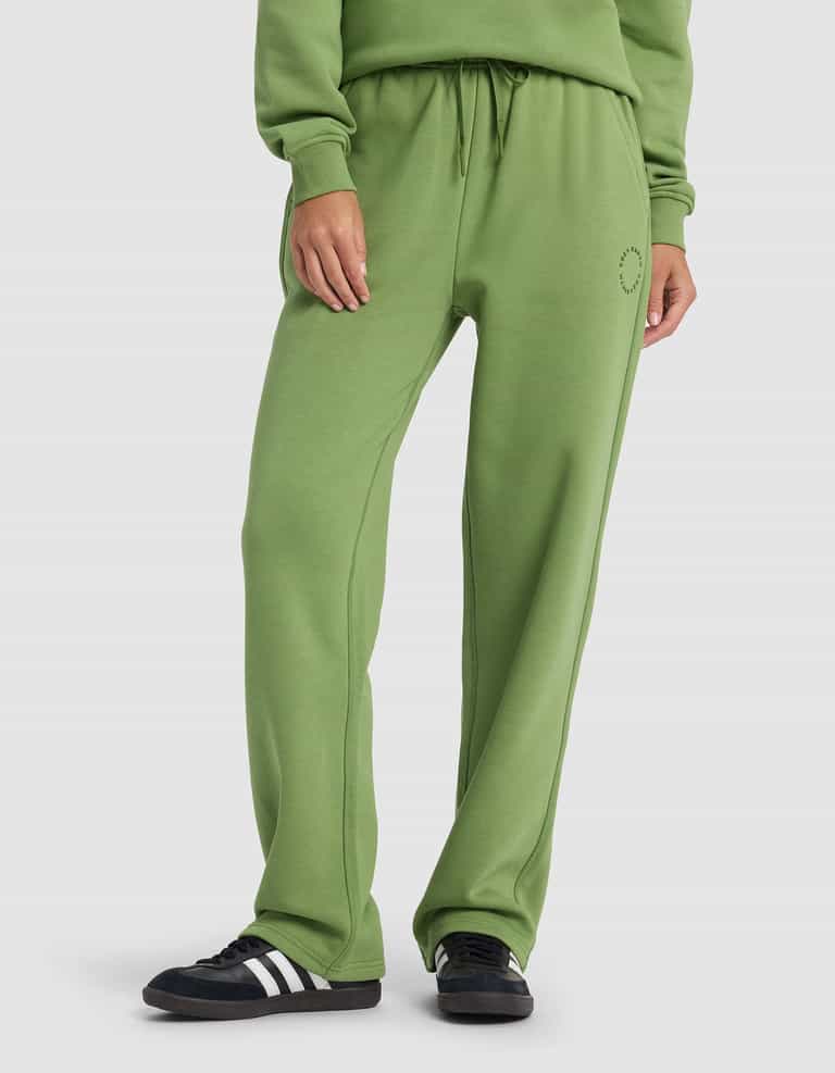 A woman wears Cozy Earth’s Women’s Cityscape 2.0 Wide Leg Pant in green with a matching sweatshirt and black sneakers with white stripes, standing against a plain white background.|Color:Clover