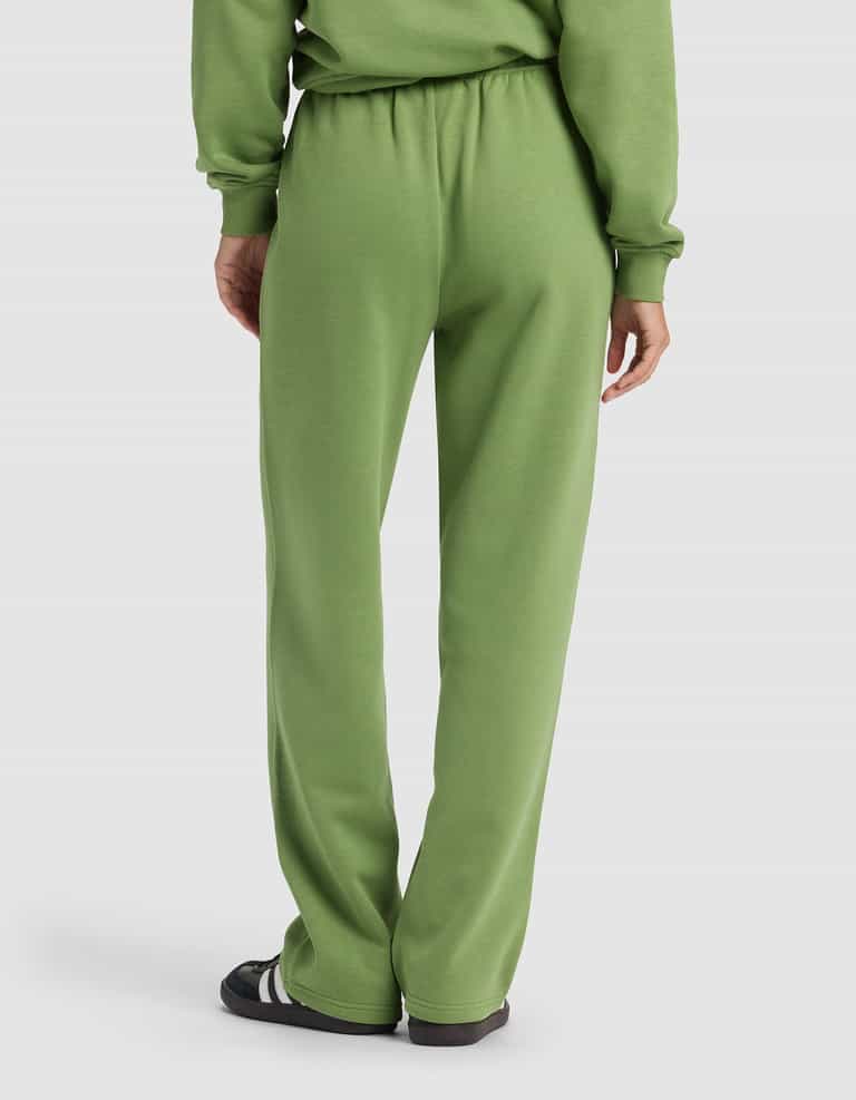 A person is seen from the back wearing the Cozy Earth Women's Cityscape 2.0 Wide Leg Pant in light green with a matching sweatshirt and black sandals with white stripes, set against a plain, light-colored background.|Color:Clover