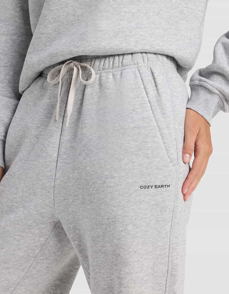 A person wears Cozy Earth Women's Cityscape 2.0 Wide Leg Pants in light gray with a drawstring waist and “COZY EARTH” branding near the pocket, paired with a matching sweatshirt and their hand in the sweatpants pocket.|Color:Heather Grey