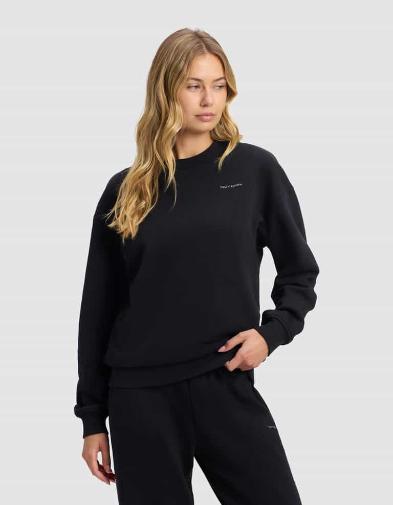 A woman with long blonde hair models the Cozy Earth Women's Cityscape 2.0 Crewneck in black, paired with matching pants, against a plain light gray background. She gazes slightly right with one hand on her hip.|Color:Jet Black
