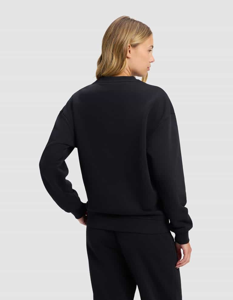 A woman with blonde hair is seen from behind wearing the Cozy Earth Women's Cityscape 2.0 Crewneck in black, paired with black pants, standing against a plain light gray background.|Color:Jet Black