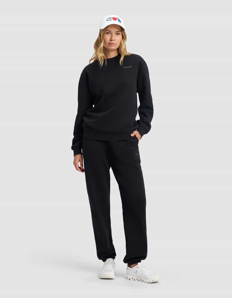A woman models the Cozy Earth Women's Cityscape 2.0 Crewneck in black, paired with matching sweatpants, a white baseball cap, and white sneakers, standing against a gray background with one hand in her pocket.|Color:Jet Black