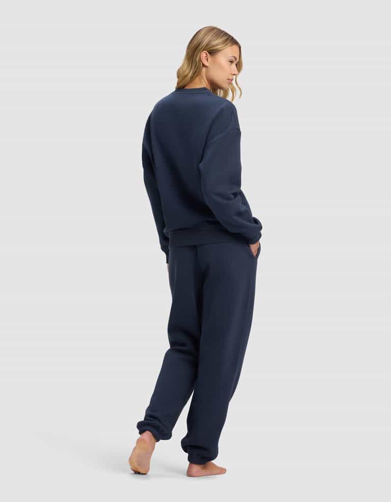 A woman stands barefoot on a light background, wearing the Cozy Earth Women's Cityscape 2.0 Crewneck & Sweatpant Set in navy blue. She faces away, glancing over her shoulder with one hand in her pocket.|Color:Eclipse