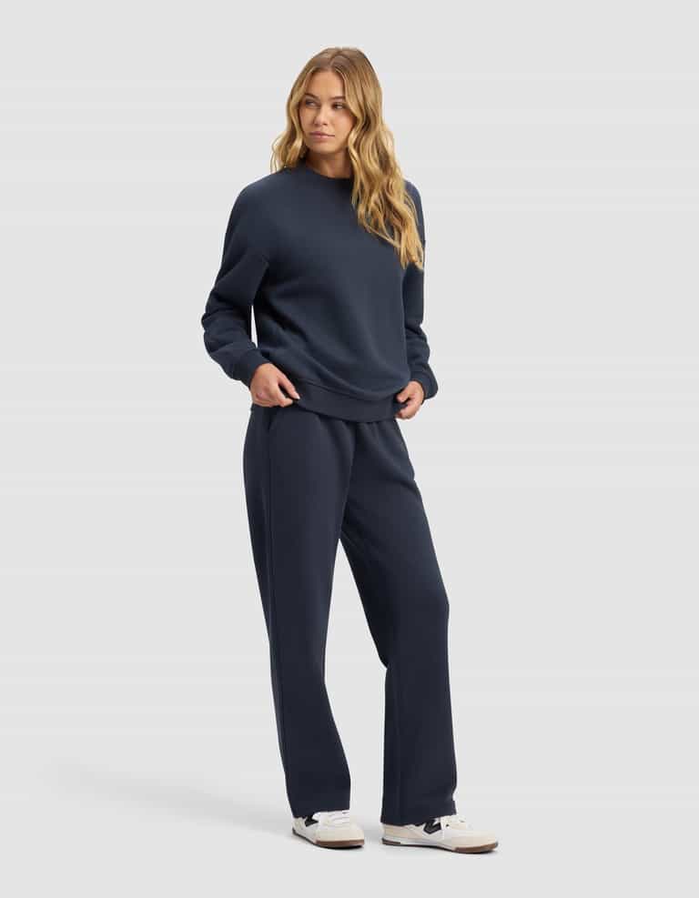 A woman with long, wavy blonde hair models the Cozy Earth Women's Cityscape 2.0 Crewneck & Wide Leg Pant Set in navy blue with white sneakers, standing against a plain light gray background.|Color:Eclipse