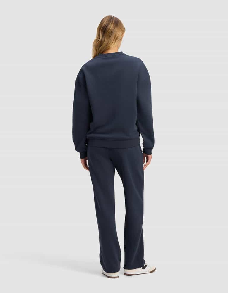 A woman with long, light brown hair stands facing away, wearing the Cozy Earth Women's Cityscape 2.0 Crewneck & Wide Leg Pant Set in navy blue with white sneakers against a plain light gray background.|Color:Eclipse