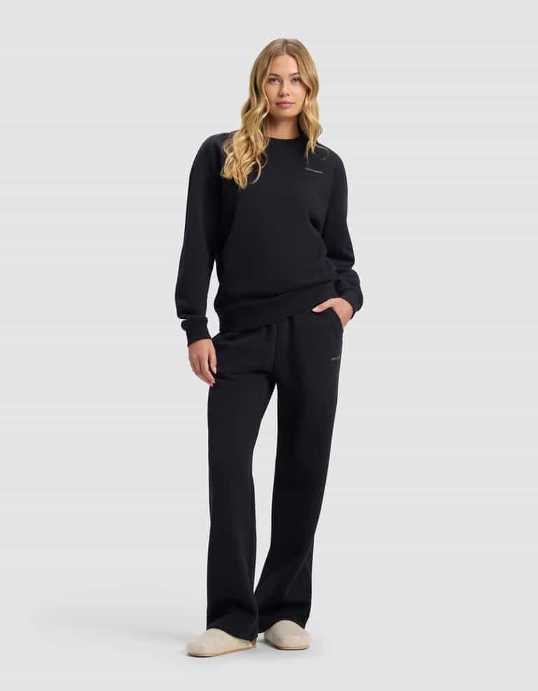 A woman with long blonde hair wears the Cozy Earth Women's Cityscape 2.0 Crewneck & Wide Leg Pant Set in black, standing forward with one hand in her pocket and light-colored slippers, against a plain light background.|Color:Jet Black