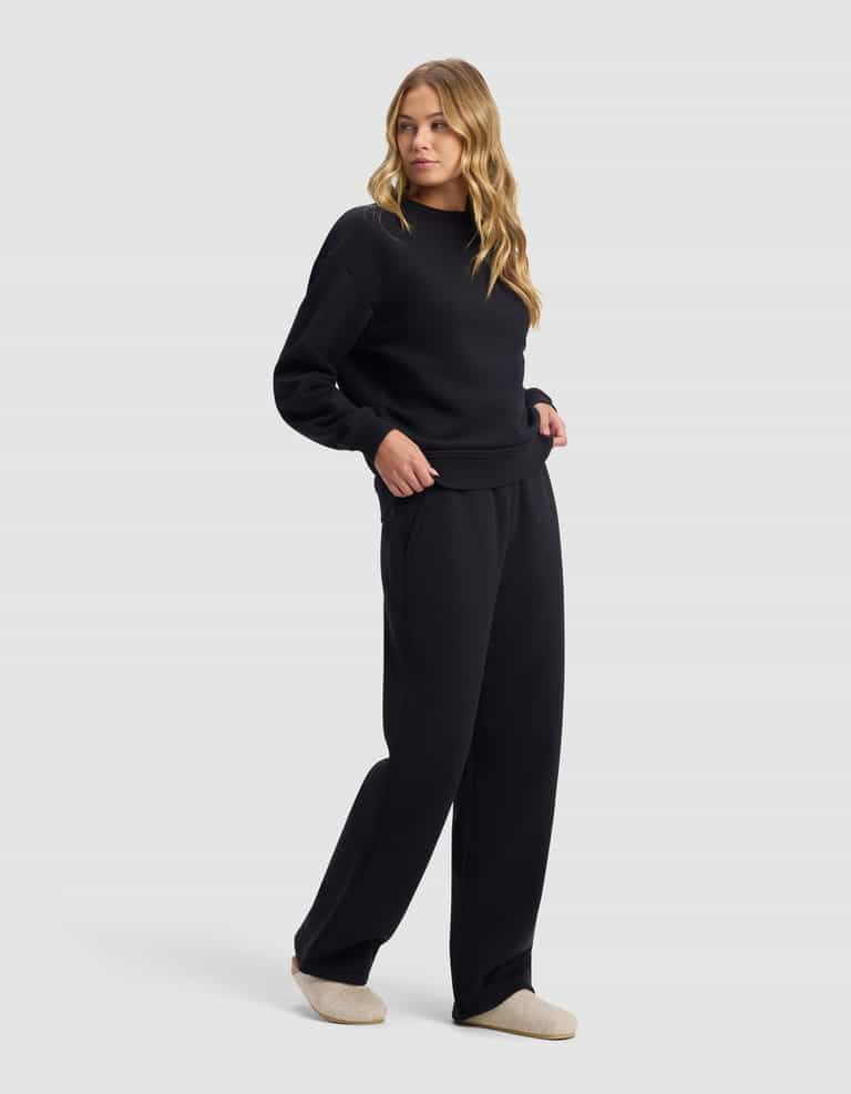 A woman with long wavy blonde hair wears the Cozy Earth Women's Cityscape 2.0 Crewneck & Wide Leg Pant Set in black with light beige slippers, standing against a plain light gray background.|Color:Jet Black