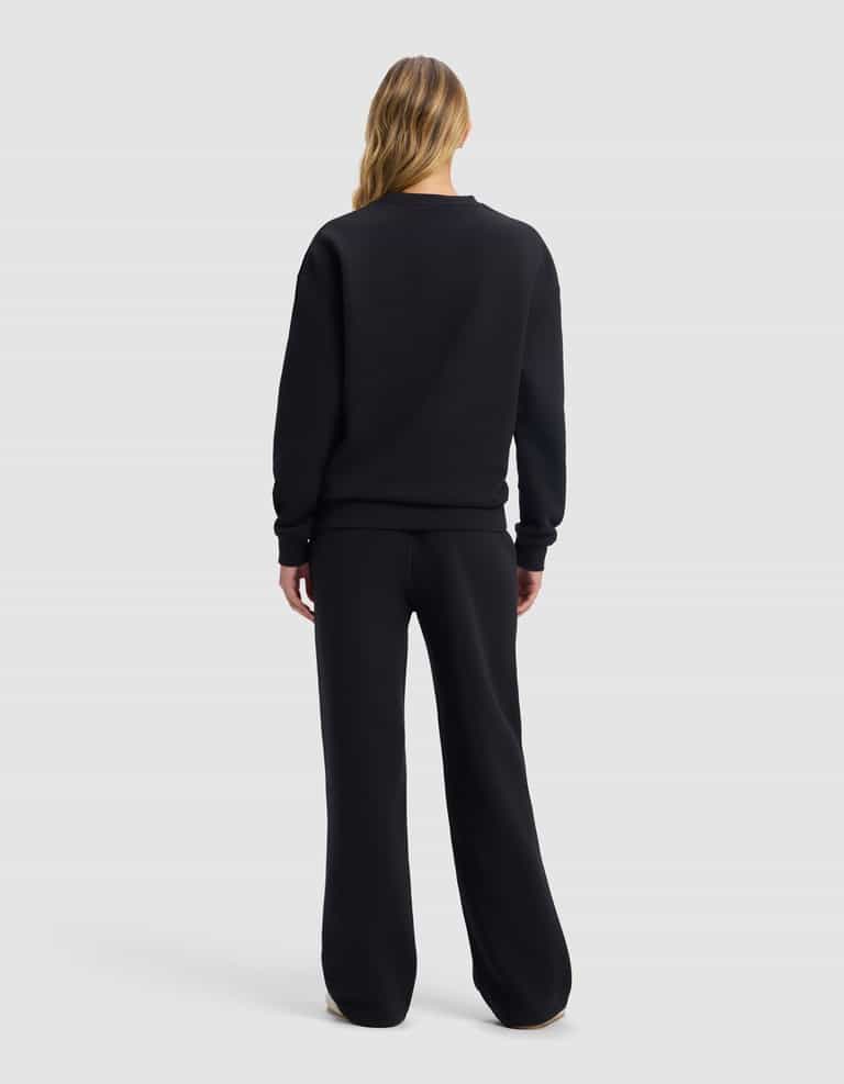 A person with long blonde hair, seen from behind, is wearing the Cozy Earth Women's Cityscape 2.0 Crewneck & Wide Leg Pant Set in black, standing against a plain light gray background.|Color:Jet Black