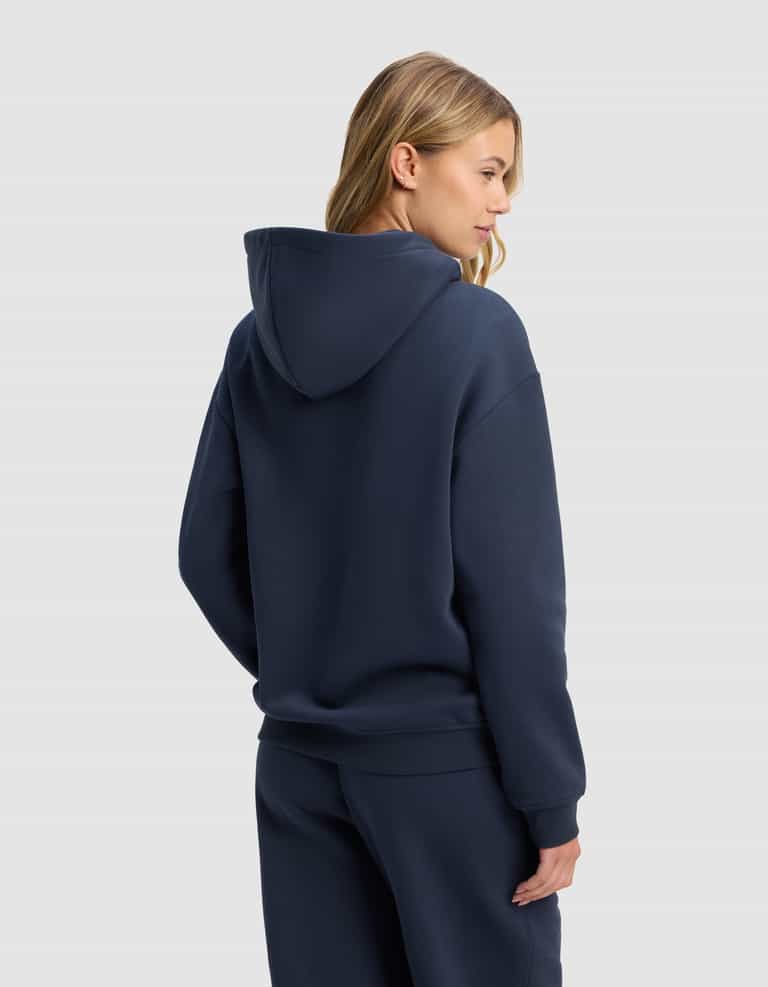 A woman stands with her back and side to the camera against a plain light gray background, wearing the Cozy Earth Women's Cityscape 2.0 Hoodie in navy blue with matching sweatpants. Her long blonde hair is visible.|Color:Eclipse