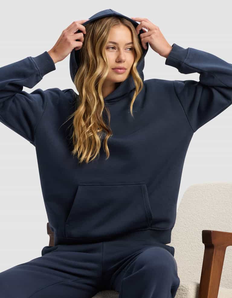 A woman with long blonde hair sits on a light chair, wearing the Cozy Earth Women's Cityscape 2.0 Hoodie in dark blue and matching sweatpants, adjusting the hood with both hands while looking slightly to the side.|Color:Eclipse