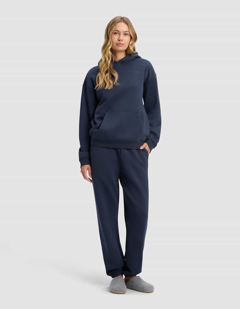 A woman with long wavy hair stands facing forward, wearing Cozy Earth’s Women’s Cityscape 2.0 Hoodie & Sweatpant Set in navy blue, paired with gray slippers, against a plain light gray background.|Color:Eclipse