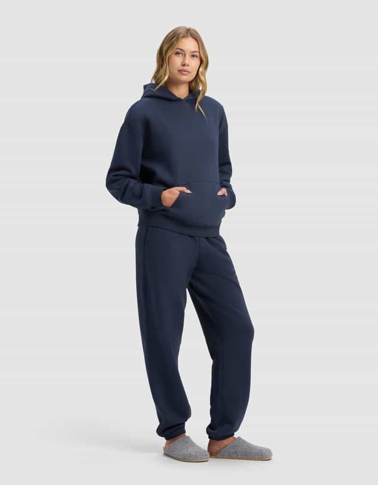 A person with long, wavy hair stands against a plain light background wearing Cozy Earth’s Women’s Cityscape 2.0 Hoodie & Sweatpant Set in navy blue, hands in the hoodie pocket, and gray slippers.|Color:Eclipse