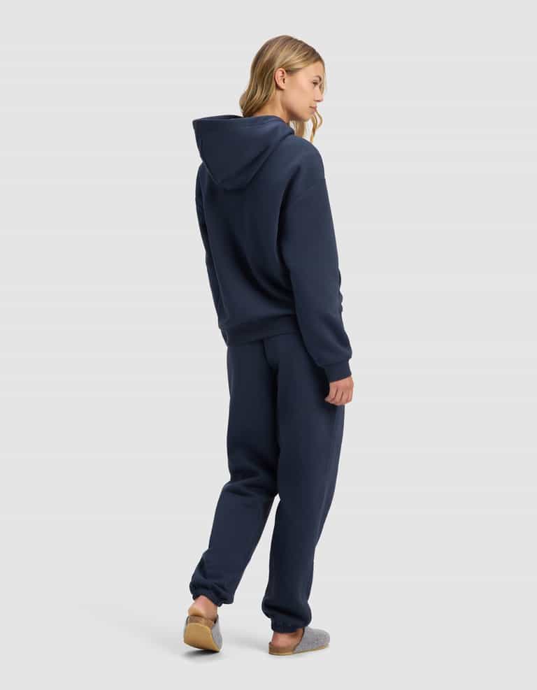 A person with long blonde hair faces away, wearing the Cozy Earth Women's Cityscape 2.0 Hoodie & Sweatpant Set in navy blue, styled with gray slippers against a plain light gray background.|Color:Eclipse