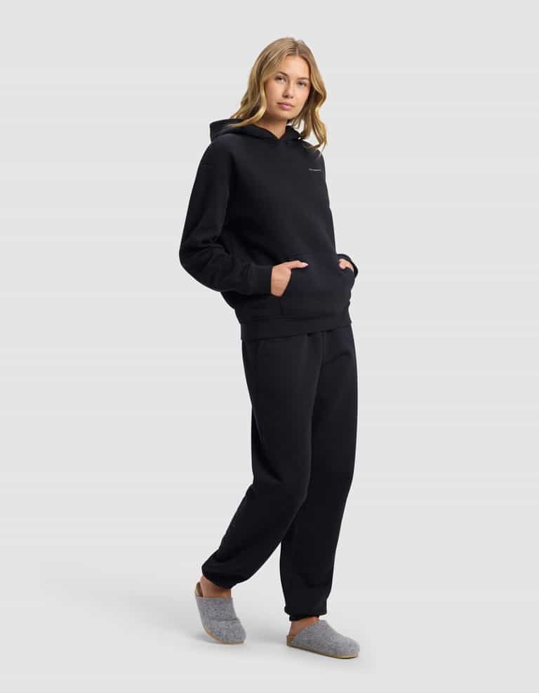 A woman with light brown hair poses against a light gray background wearing Cozy Earth's Women's Cityscape 2.0 Hoodie & Sweatpant Set in black, with her hands in the front hoodie pocket and gray slippers on her feet.|Color:Jet Black
