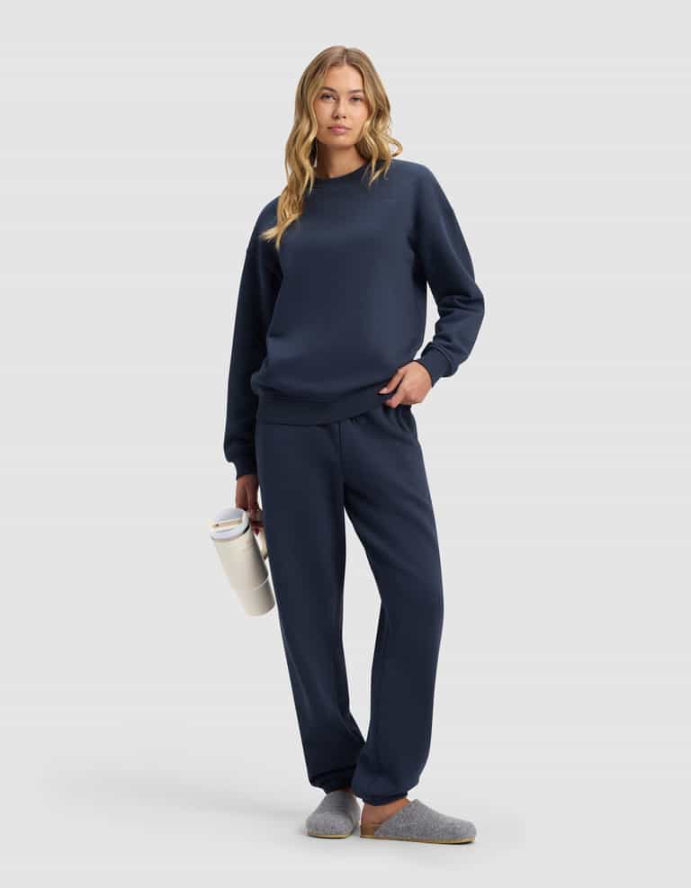 A woman in Cozy Earth's Women's Cityscape 2.0 Sweatpant, paired with a navy blue sweatshirt and gray slippers, holds a white water bottle. She has long wavy blonde hair, a relaxed look, and stands against a plain light gray background.|Color:Eclipse
