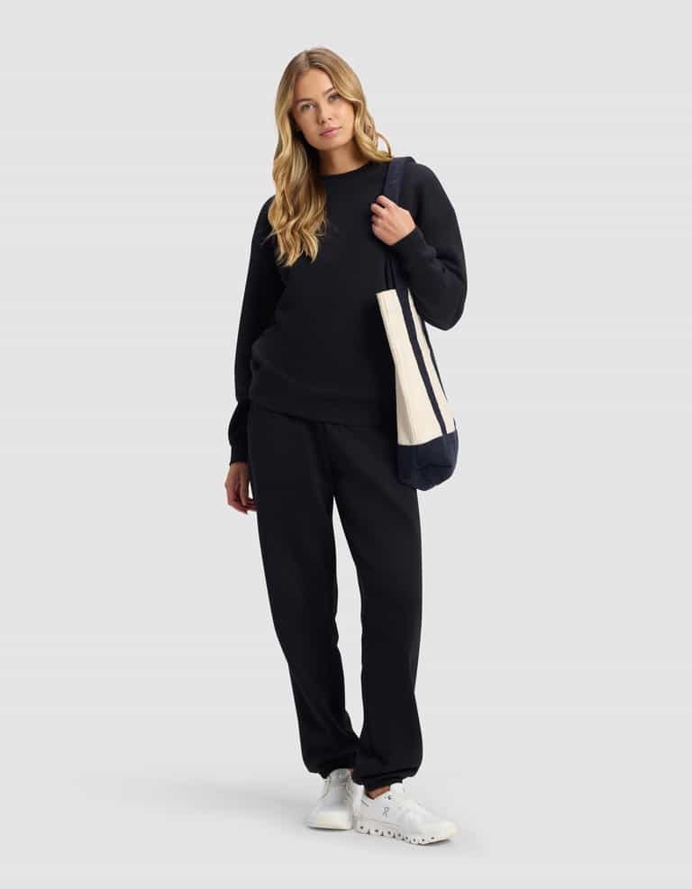 A woman with long blonde hair wears the Cozy Earth Women's Cityscape 2.0 Sweatpant in black, a matching sweatshirt, white sneakers, and carries a beige tote bag, standing against a plain light gray background.|Color:Jet Black