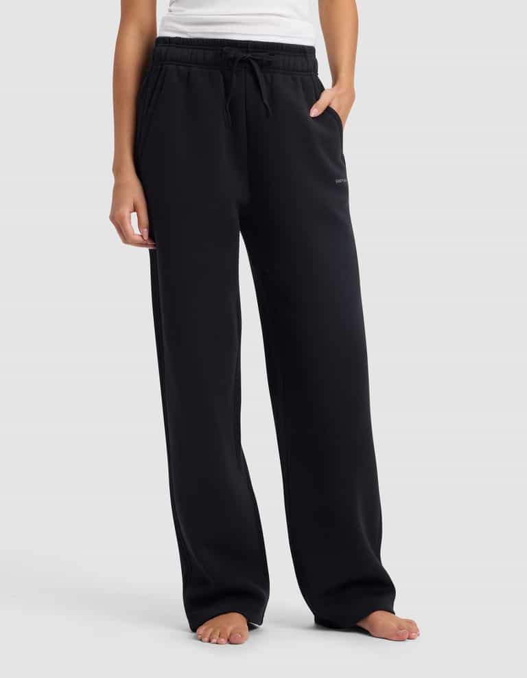 A person stands barefoot in Cozy Earth's Women's Cityscape 2.0 Wide Leg Pant, a loose-fitting black sweatpant, paired with a white sleeveless top, one hand in their pocket, against a plain light gray background.|Color:Jet Black