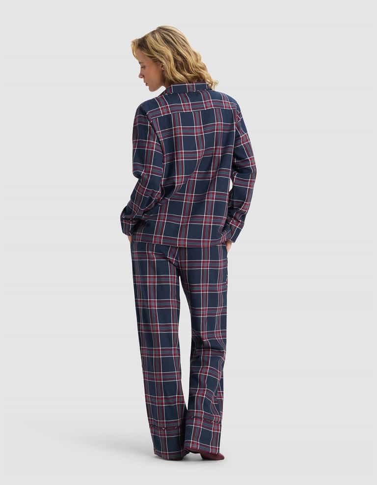 A woman, hands in pockets, wears the Cozy Earth Women's Farmhouse Long Sleeve Pajama Set in navy blue, red, and white plaid with long sleeves and pants, standing against a plain light gray background.|Color:Eclipse Plaid