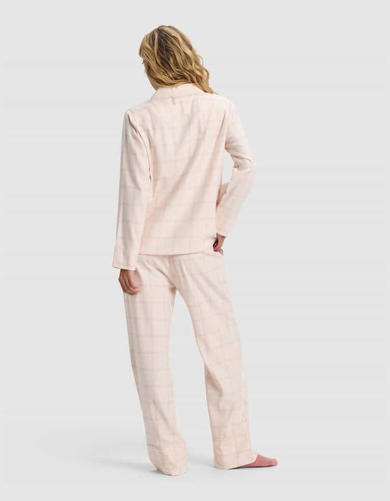 A woman with wavy blonde hair stands with her back to the camera, wearing Cozy Earth's Women's Farmhouse Long Sleeve Pajama Set in light pink plaid and pink slippers against a plain light gray background.|Color:Quartz Plaid