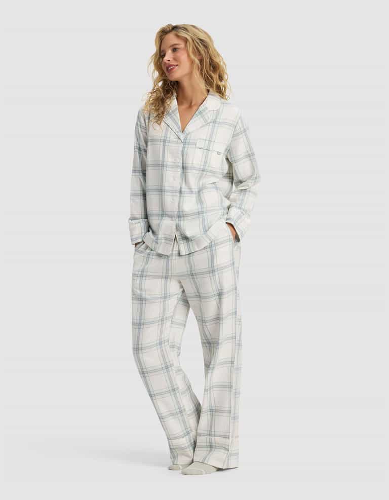 A woman with wavy blonde hair smiles and looks to the side, hands in her pockets, wearing the Cozy Earth Women's Farmhouse Long Sleeve Pajama Set—a loose-fitting, button-up plaid ensemble in light colors.|Color:Wintergreen Plaid