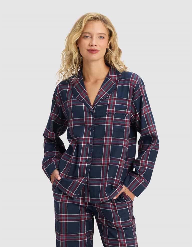 A woman with wavy blonde hair wears the Cozy Earth Women's Farmhouse Long Sleeve Pajama Top in a navy blue plaid with red and white stripes, standing with her hands in her pockets against a plain light gray background.|Color:Eclipse Plaid