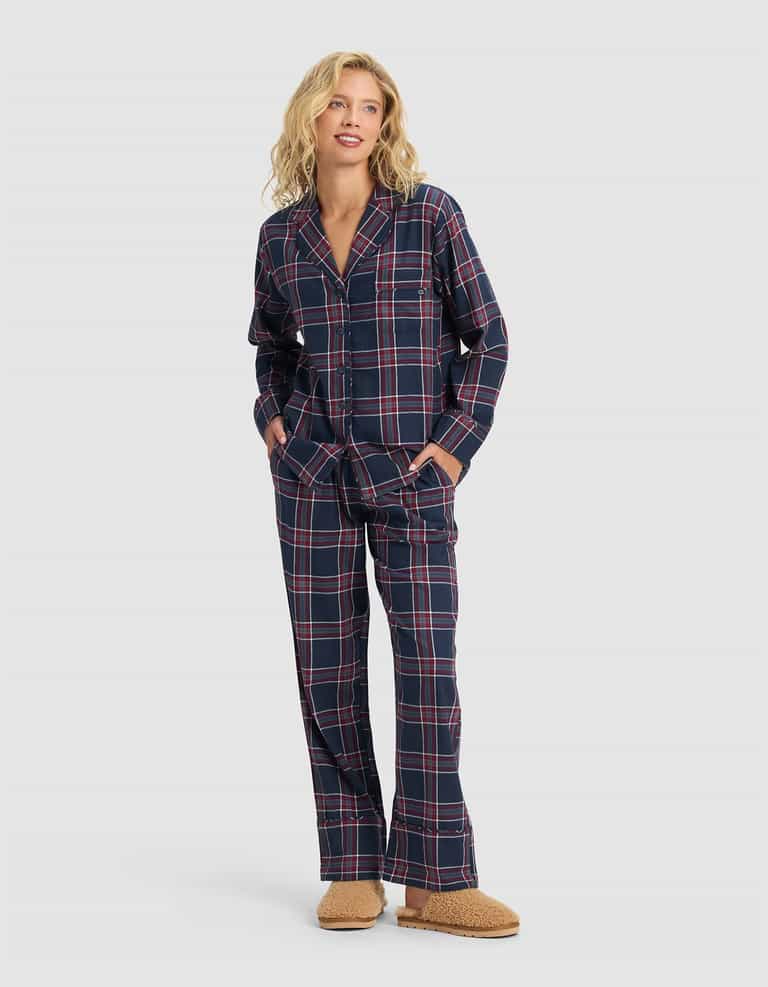 A person stands smiling, wearing the Cozy Earth Women's Farmhouse Long Sleeve Pajama Top in navy blue with red and white plaid, paired with loose pajama pants and tan fuzzy slippers. The top features buttons and pockets.|Color:Eclipse Plaid