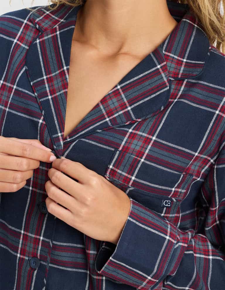 A person is buttoning up the Cozy Earth Women's Farmhouse Long Sleeve Pajama Top in a navy blue, red, and white plaid pattern, with only their torso and hands visible.|Color:Eclipse Plaid