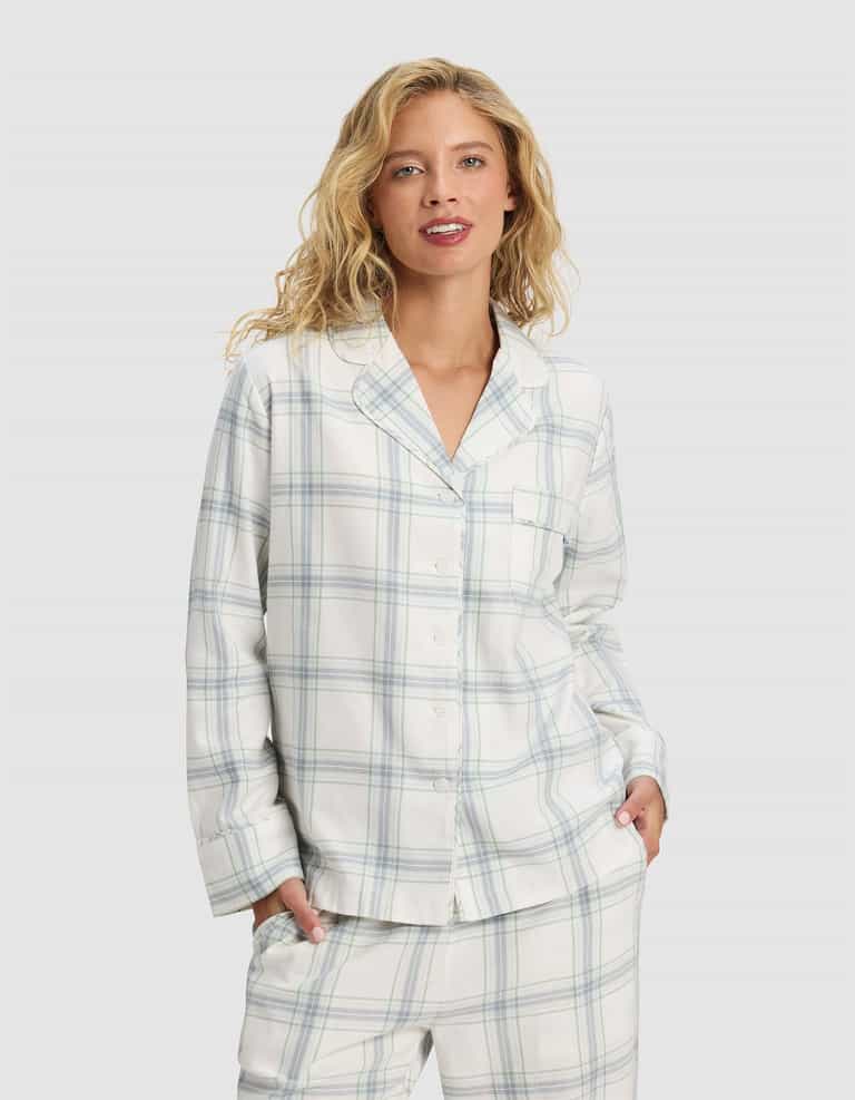 A woman with wavy blonde hair wears the Cozy Earth Women's Farmhouse Long Sleeve Pajama Top in a white fabric with light blue and gray plaid, standing hands-in-pockets against a plain light background.|Color:Wintergreen Plaid