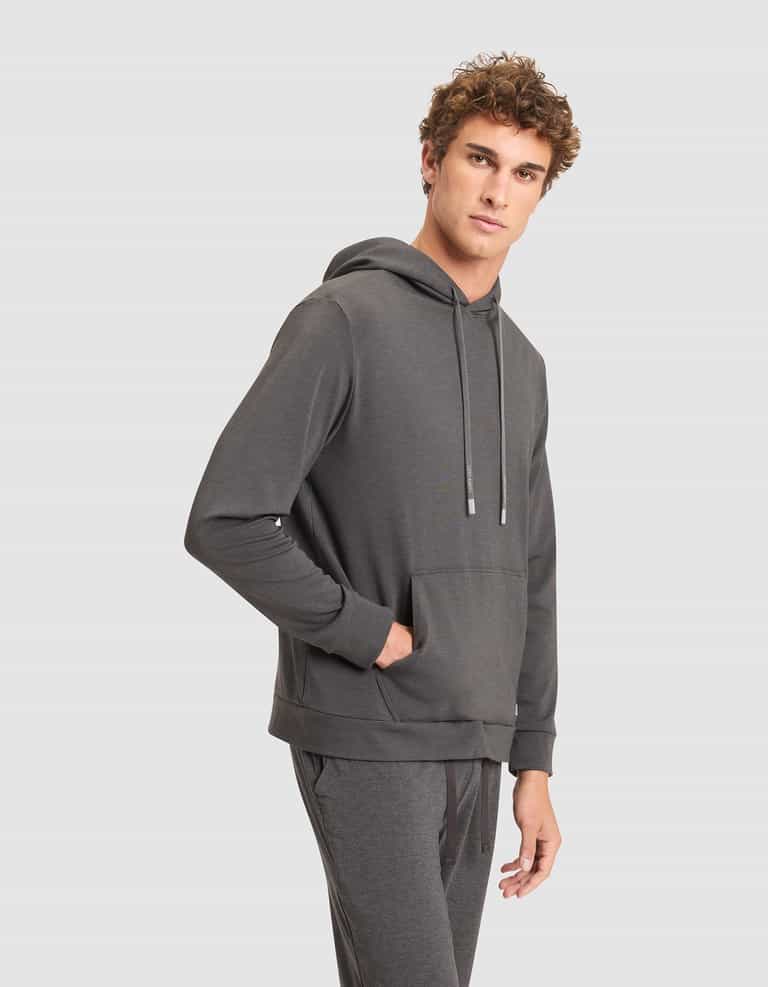 A young man with curly hair models the Cozy Earth Men's Brushed Bamboo Hoodie in dark gray, paired with matching joggers, standing with one hand in his pocket against a plain light gray background. |Color:Charcoal