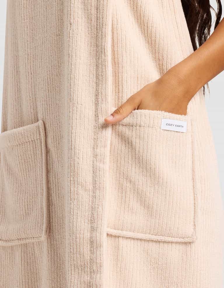 Someone wears Cozy Earth's Ribbed Terry Bath Wrap in beige, featuring ribbed fabric and large front pockets—one hand rests inside a pocket with the Cozy Earth label showing.|Color:Petal