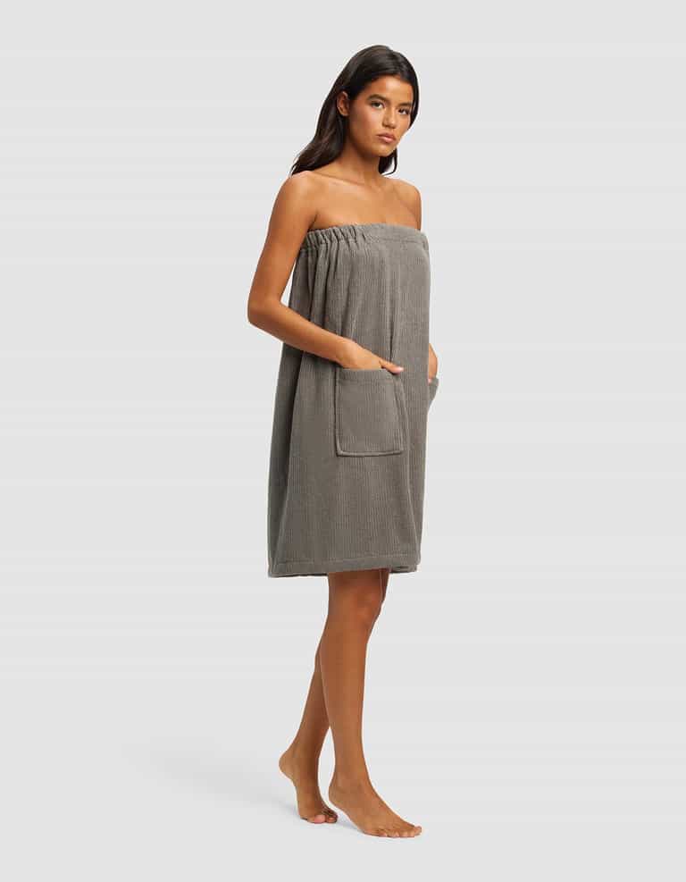 A woman stands barefoot against a plain background, wearing the Cozy Earth Ribbed Terry Bath Wrap in grey. Its strapless design features pockets, and she rests her hands in them. Her straight dark hair frames her neutral expression.|Color:Charcoal