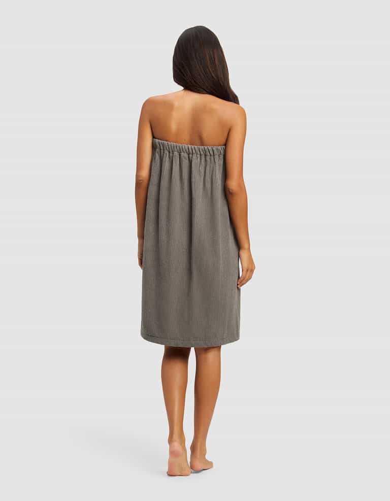 A woman with long brown hair stands barefoot, facing away from the camera, wearing the Cozy Earth Ribbed Terry Bath Wrap—a gray, strapless, knee-length towel dress—against a plain light background.|Color:Charcoal