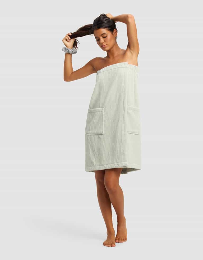 A woman stands barefoot against a plain background, adjusting her ponytail while wearing the Cozy Earth Ribbed Terry Bath Wrap in light green with two front pockets and a matching scrunchie on her wrist.|Color:Laurel