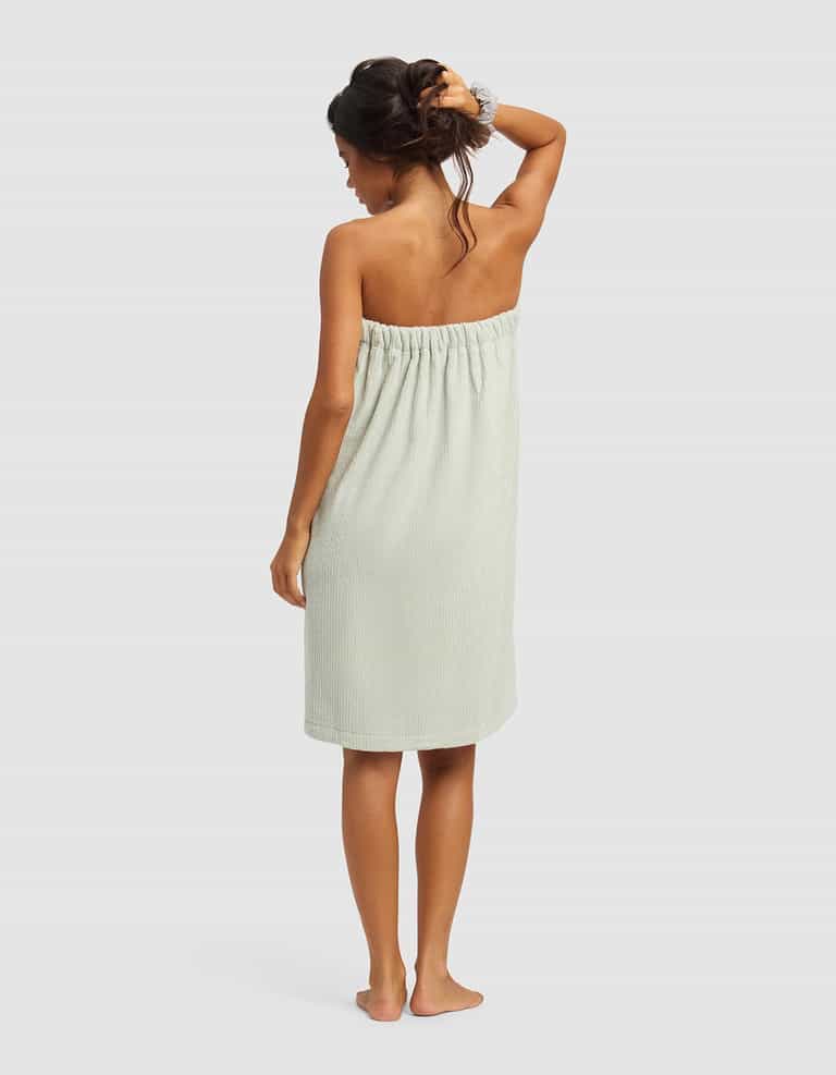 A woman stands barefoot with her back to the camera, wearing the Cozy Earth Ribbed Terry Bath Wrap in light green and a hair scrunchie, one arm raised to touch her hair, set against a plain light background.|Color:Laurel